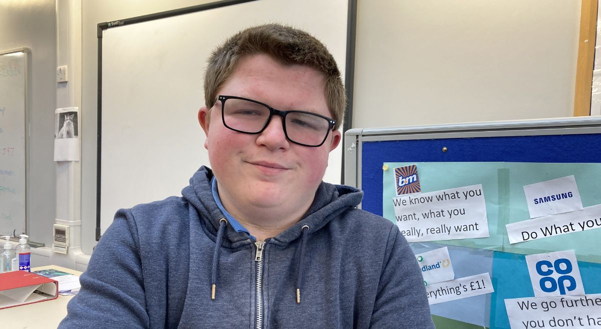 Young man, Gerard Blaney, with short dark hair, wearing glasses and a dark blue hoodie pictured in front of a notice board in a classroom setting.